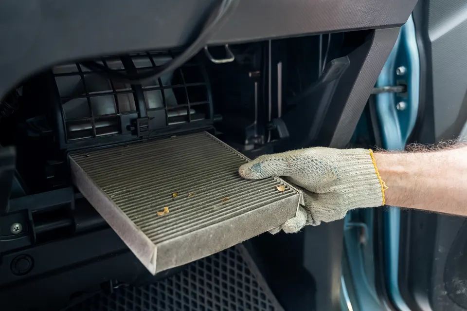 A gloved hand removing a dirty cabin air filter from behind the glove box