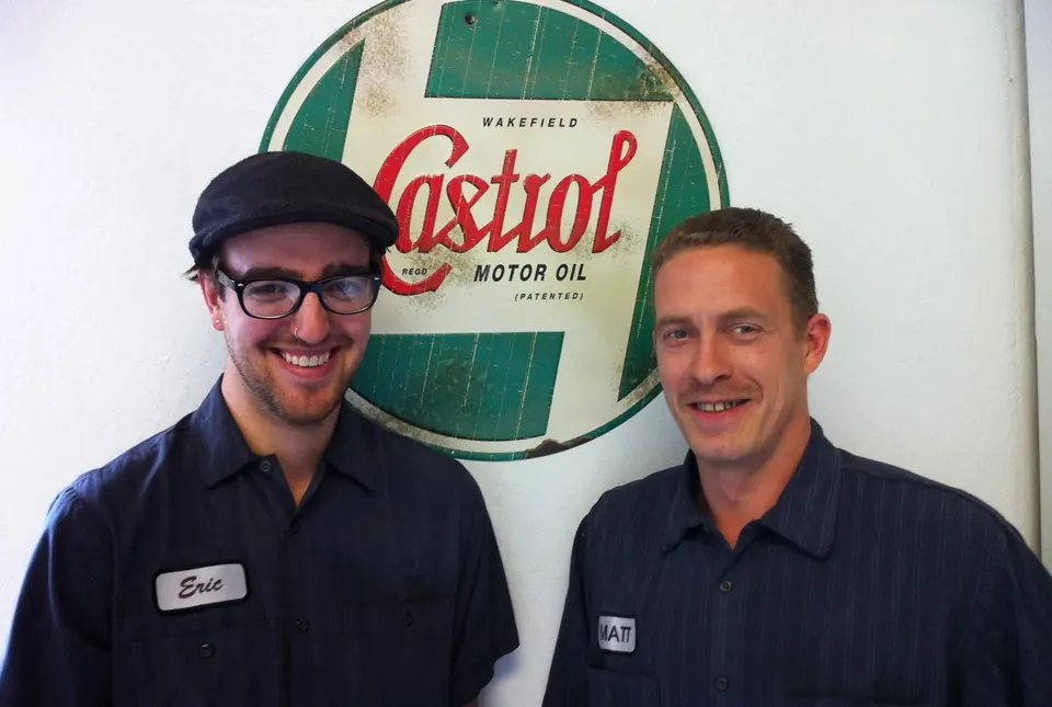 Two Xpress technicians smiling in front of a vintage motor oil sign at the Reno Northwest service center
